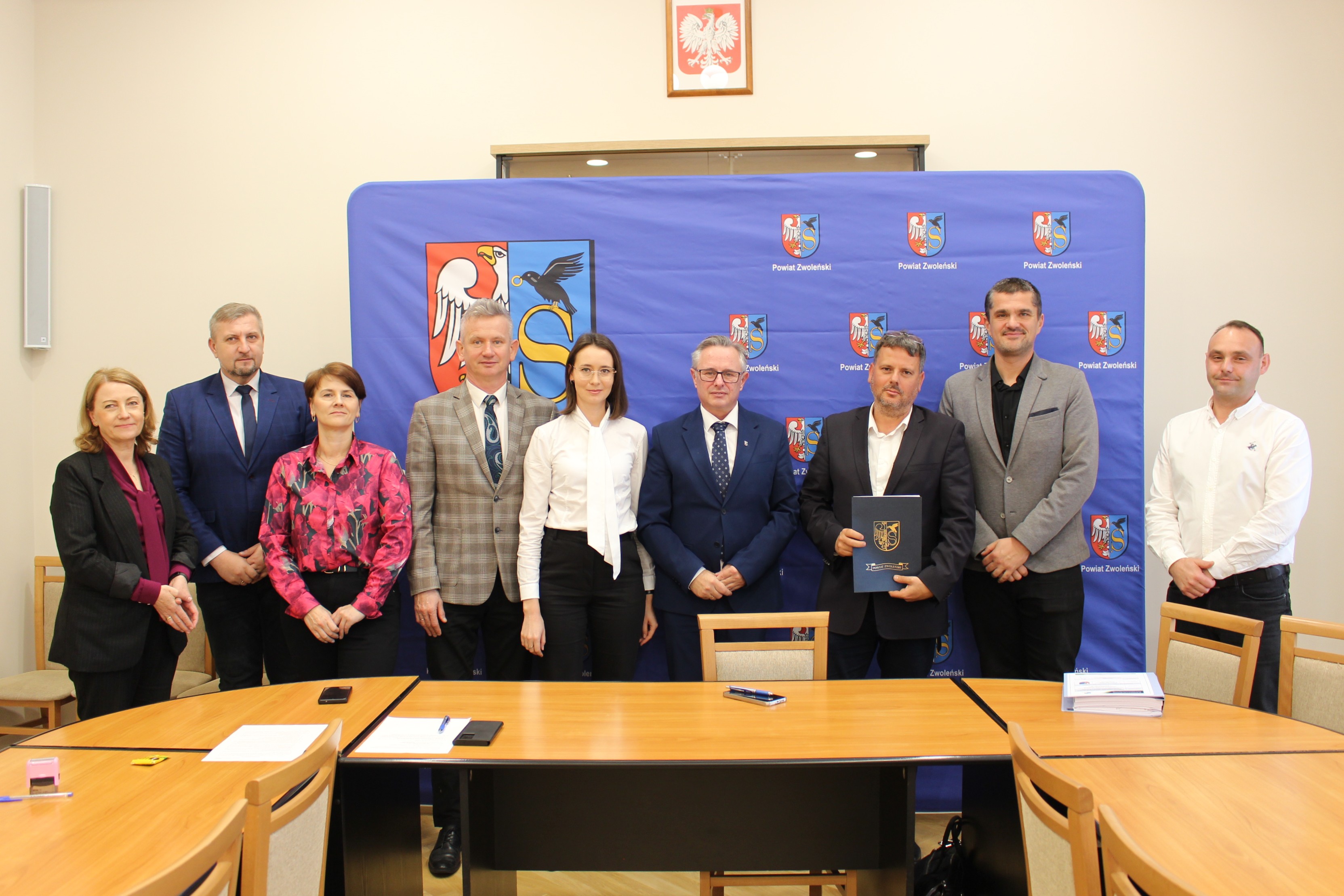 Zdjęcie przedstawia grupę osób stojących na tle ścianki promocyjne Powiatu Zwoleńskiego na sali narad w Starostwie Powiatowym w Zwoleniu.