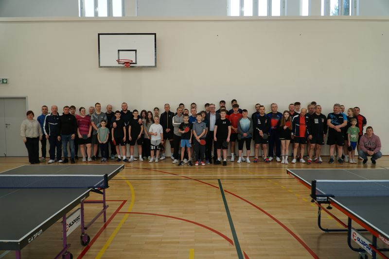 Zdjęcie przedstawia grupę os&oacute;b stojących na sali gimnastycznej podczas turnieju tenisa stołowego.