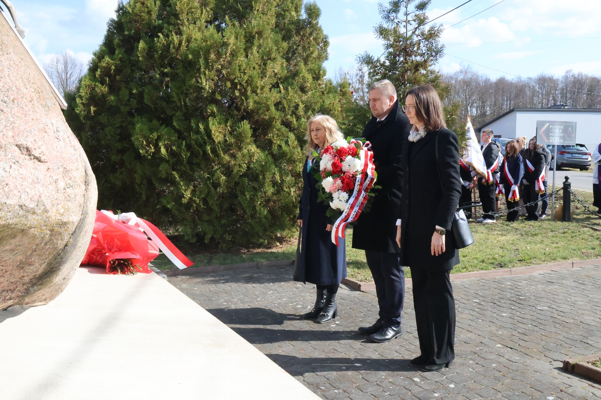 Zdjęcie przedstawia troje ludzi składających wiązankę pod pomnikiem w Karolinie.