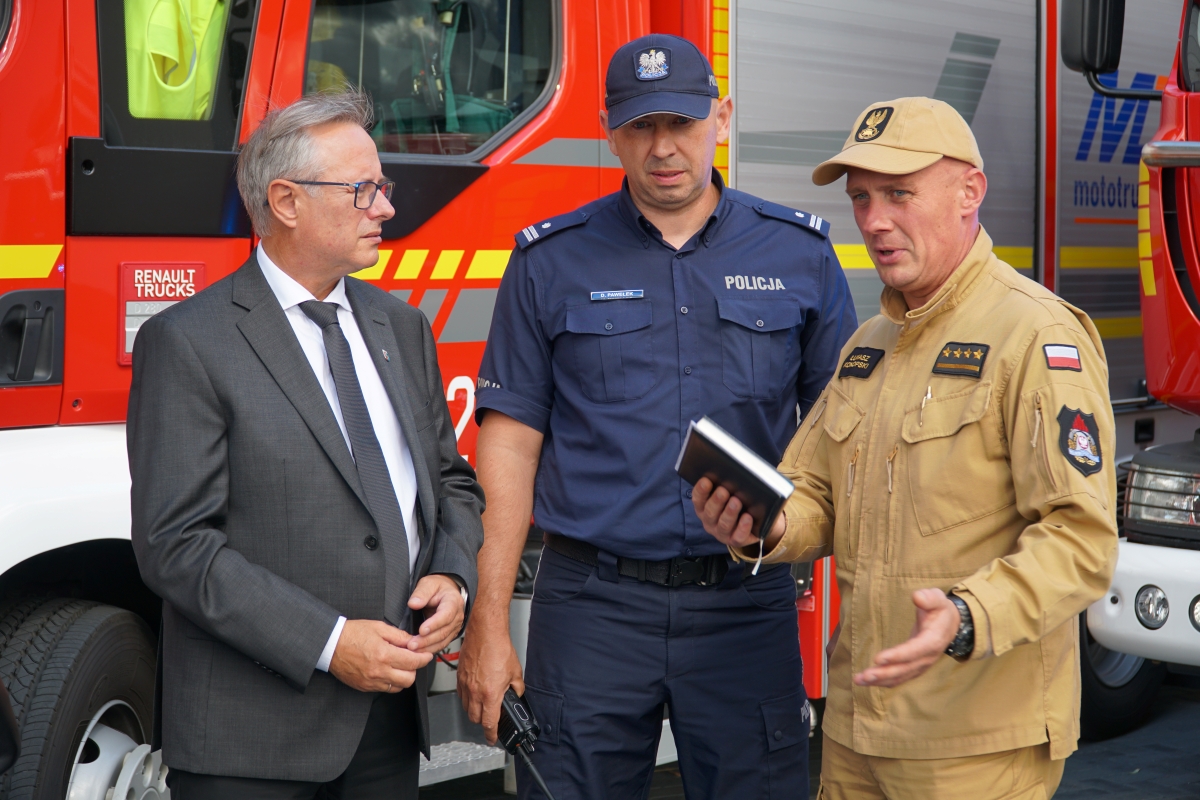 Zdjęcie przedstawia trzech mężczyzn stojących na tle radiowozu strażackiego.
